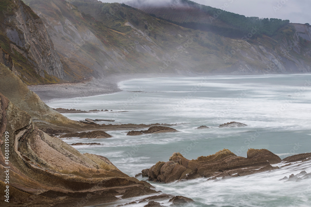 Fototapeta premium Powerful waves crashing against rugged flysch cliffs at Itzurun Beach, Cantabria, generating dramatic moody seascape under overcast northern Spanish skies