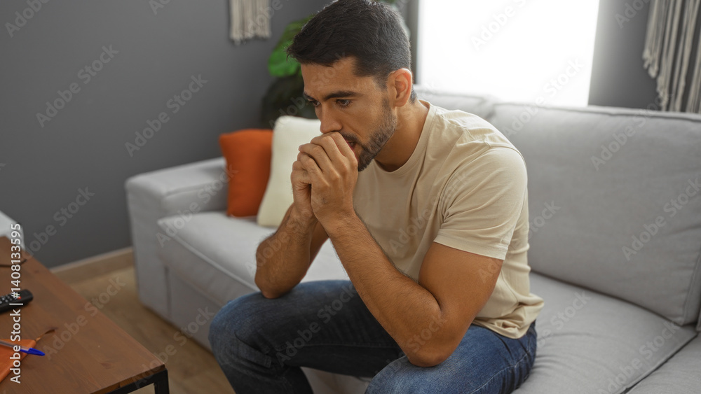 Handsome young man with beard in jeans and t-shirt sitting thoughtfully on a sofa in a modern living room filled with natural light Handsome young man with beard in jeans and t-shirt sitting thoughtfully on a sofa in a modern living room filled with natural light