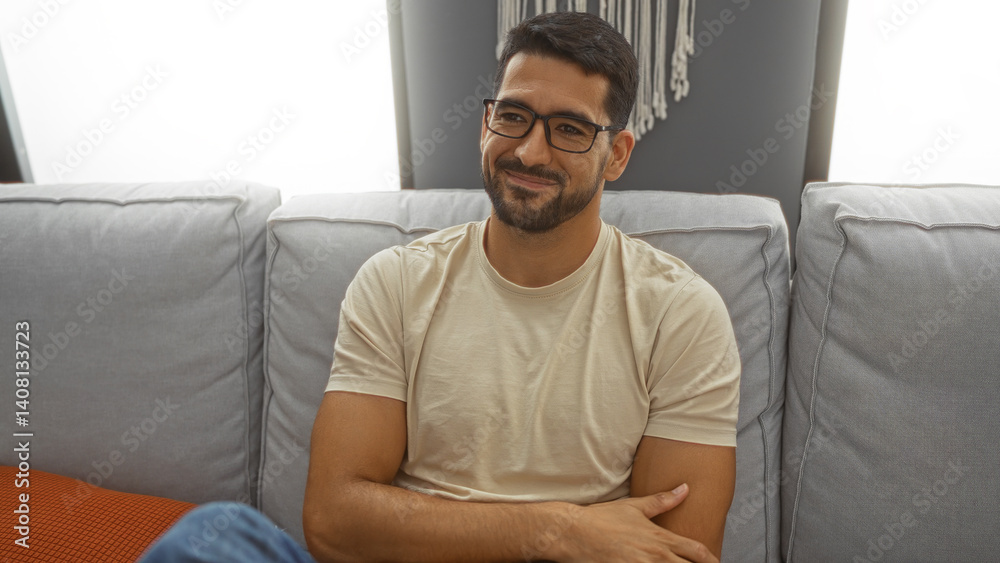 Young man with beard sitting on a sofa with arms crossed, indoors, in a bright living room, wearing glasses, looking content, in a modern home setting. Young man with beard sitting on a sofa with arms crossed, indoors, in a bright living room, wearing glasses, looking content, in a modern home setting.