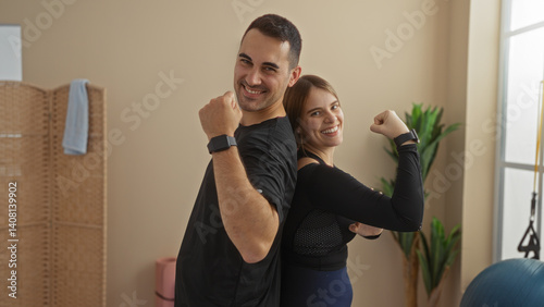 Obraz na płótnie Man and woman smiling while making strong arm gestures standing back to back in a gym indoor setting showcasing fitness, happiness, and partnership. w salonie