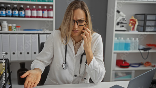 Woman doctor talking on phone in clinic while sitting at desk with medical equipment in background showcasing a busy hospital environment.