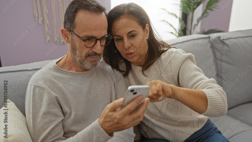 Fototapeta premium Hispanic couple sitting together on a sofa in their living room, smiling and looking at a smartphone, depicting a warm family moment indoors.