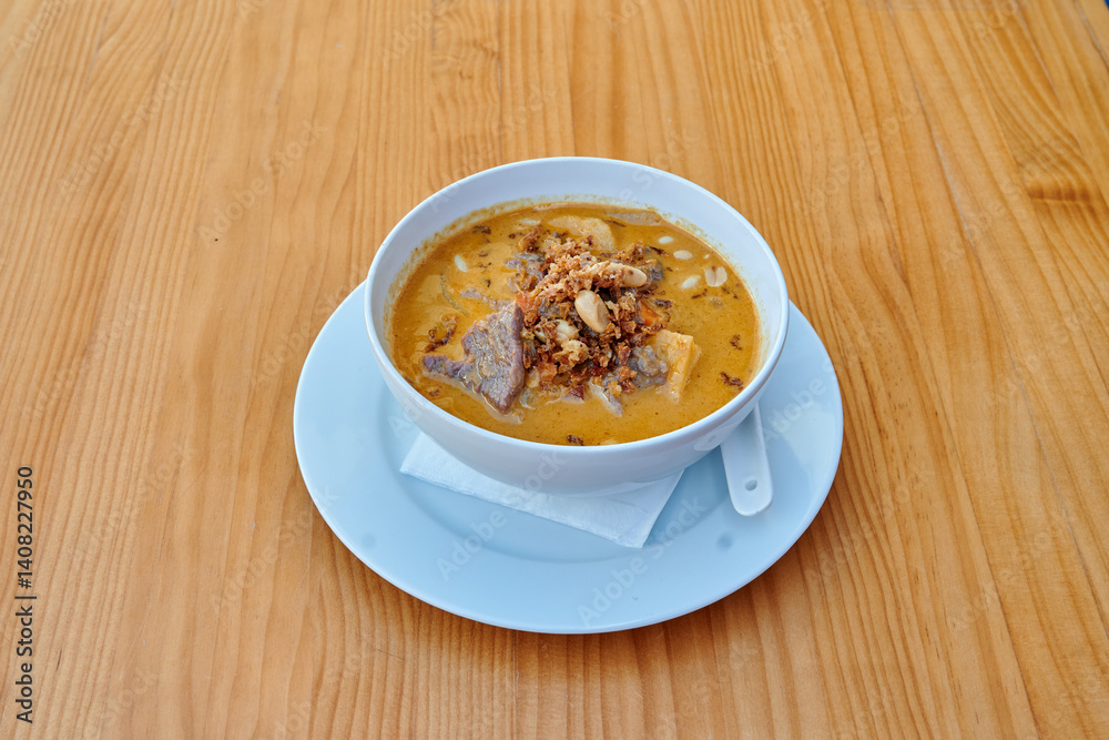 A bowl of soup with meat and nuts on a white plate. The bowl is on a wooden table