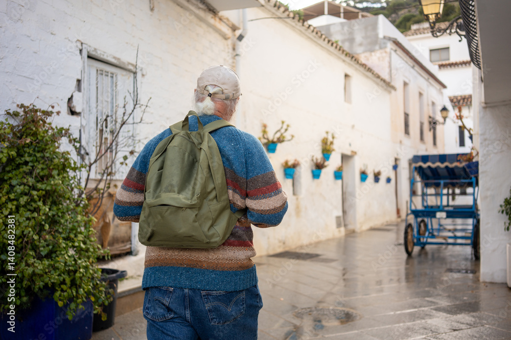 Senior man with gray hair and green backpack walking through a narrow Spanish street with white walls, blue flower pots, and a vintage cart under cloudy weather after rain. Senior man with gray hair and green backpack walking through a narrow Spanish street with white walls, blue flower pots, and a vintage cart under cloudy weather after rain.