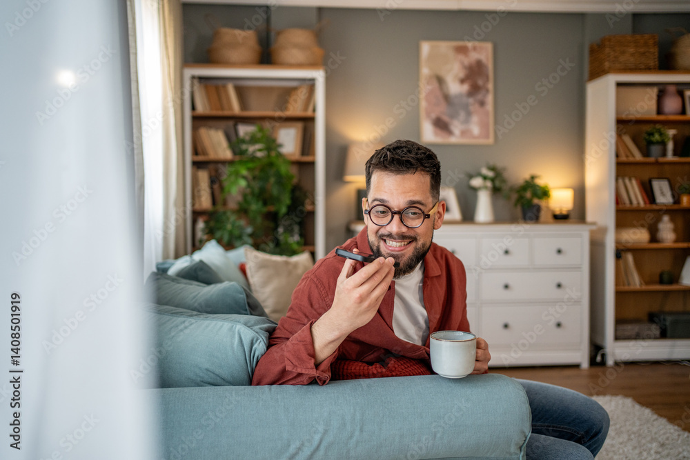 Young man recording voice message while drinking coffee on sofa at home Young man recording voice message while drinking coffee on sofa at home