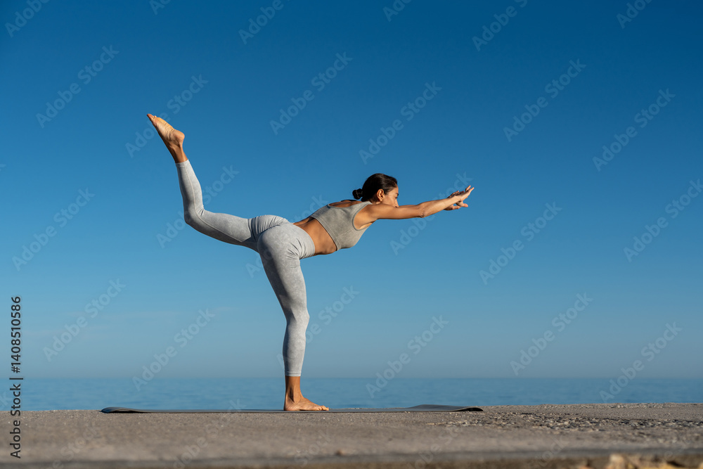 Obraz premium Woman practicing yoga balance pose on a sunny day at the beach with a bright blue sky in the background, showcasing strength and flexibility.