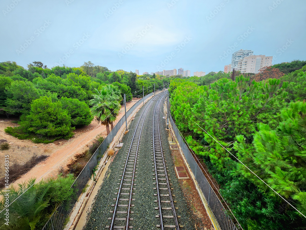 Obraz premium Curving railway tracks pass through a lush green forest with city buildings in the background, blending infrastructure with nature under an overcast sky.