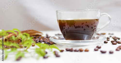 Transparent cup filled with steaming coffee sits on a white table, surrounded by scattered coffee beans, cinnamon sticks, and delicate green leaves, creating a cozy and inviting scene