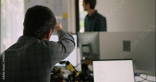 Engineer raises a point during a discussion with a teammate while surrounded by monitors, laptops, and robotic components in the lab.