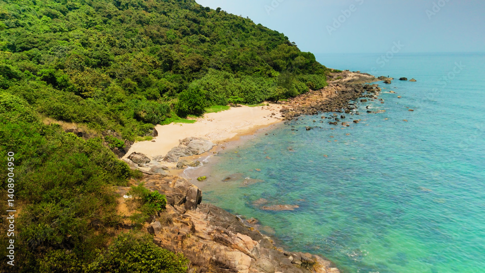 Fototapeta premium Lush tropical vegetation covering a hill sloping down to a sandy beach and transparent ocean water with rocks at the coastline inviting to swim and relax