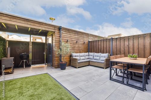 A stylish outdoor patio featuring comfortable seating, a dining table, and a BBQ grill. The space is complemented by greenery and a clean modern design.