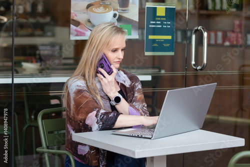 Attractive mid-age woman with heterochromia (different eye colors) working on a laptop at an outdoor cafe table, talking on her phone, seated comfortably in a casual setting in a modern coffee shop
