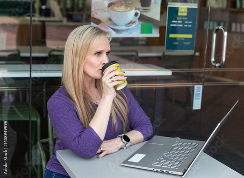 Attractive mid-age woman with heterochromia (different eye colors) working on a laptop at an outdoor cafe table, seated comfortably in a casual setting with a modern coffee shop background
