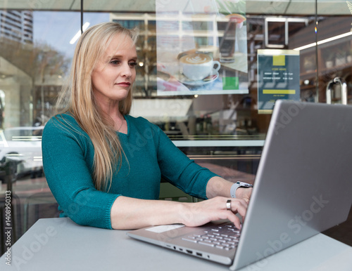 Attractive mid-age woman with heterochromia (different eye colors) working on a laptop at an outdoor cafe table, seated comfortably in a casual setting with a modern coffee shop background