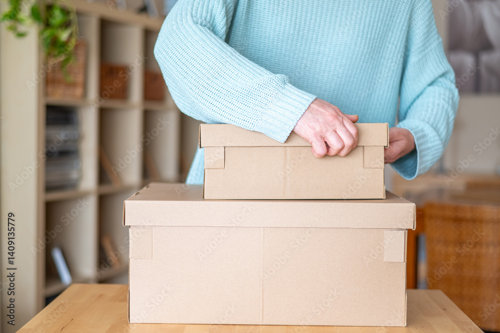 Woman is carefully placing a cardboard box on top of another, organizing her belongings in her new house, surrounded by shelves and a wooden table Woman is carefully placing a cardboard box on top of another, organizing her belongings in her new house, surrounded by shelves and a wooden table