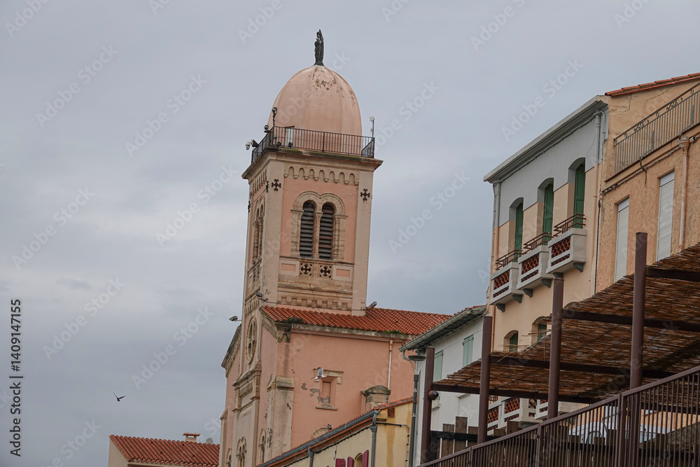 A beautiful church tower rises above colorful buildings in a quaint town, captured on a cloudy day. The unique architecture reflects local heritage and culture. Porte-Vendre, France