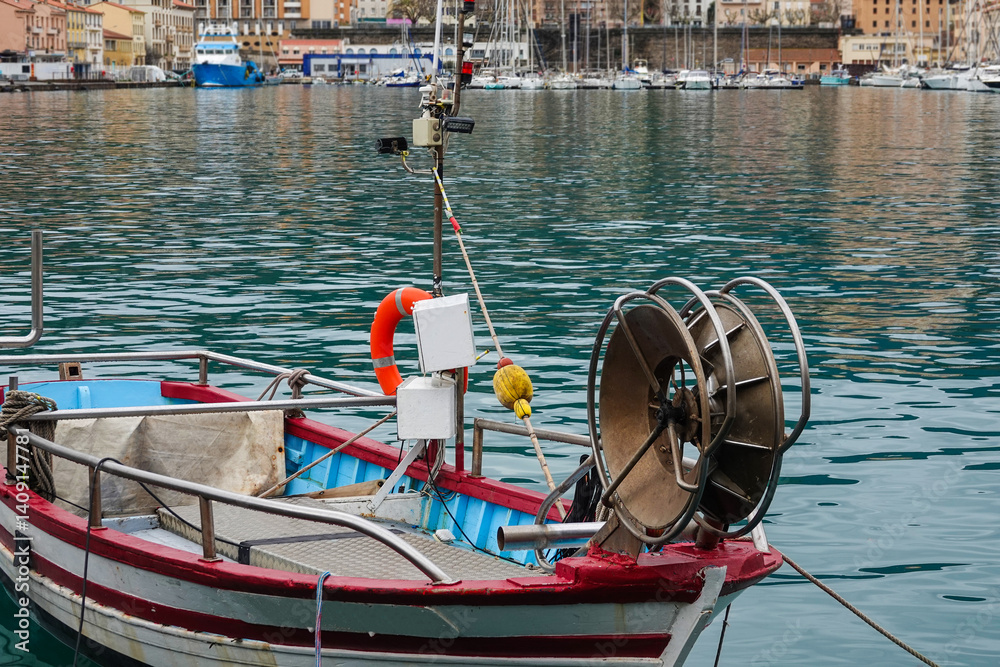 A small fishing boat is anchored in a serene harbor, surrounded by colorful buildings and larger yachts.