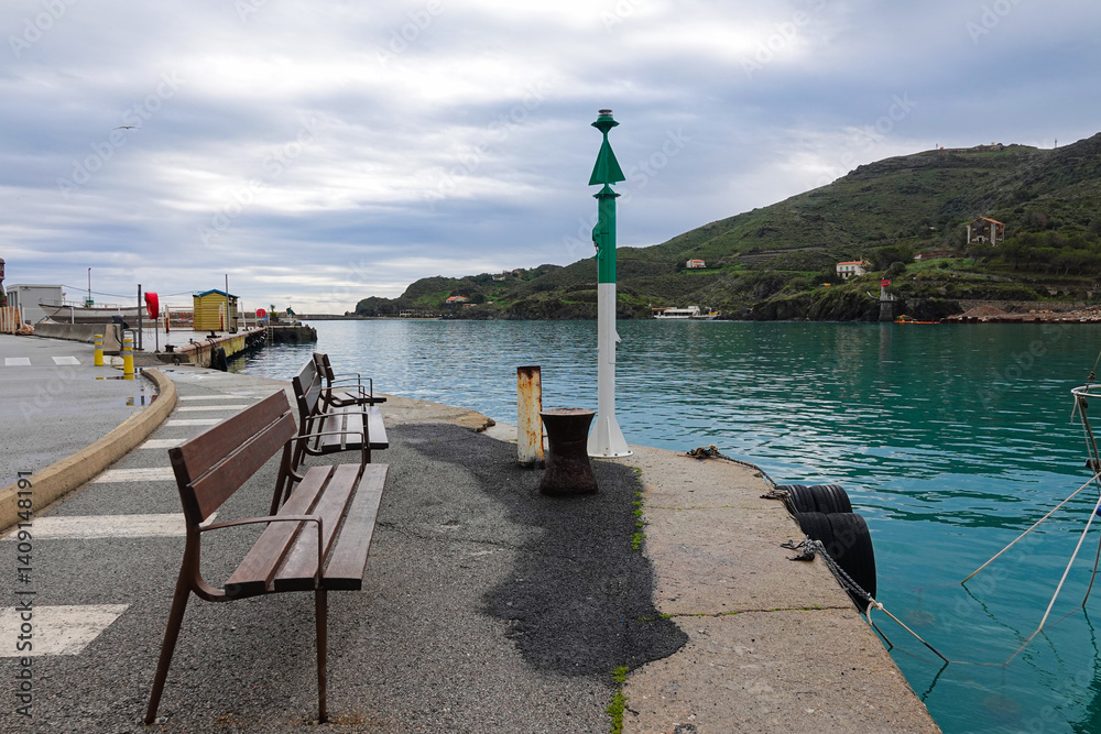 Benches line the edge of a tranquil harbor, where still waters reflect gray skies.