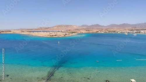 Aerial view of a crystal-clear blue bay bordered by desert hills, with boats, a sailboarder, and distant seaside town under a clear sky