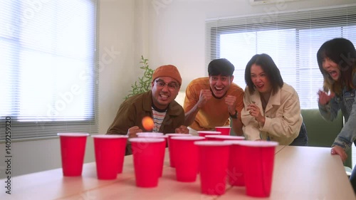 Happy asian diverse group friends playing together with beer pong at home office standing at the table enjoying time together. Office leisure and board games concept.