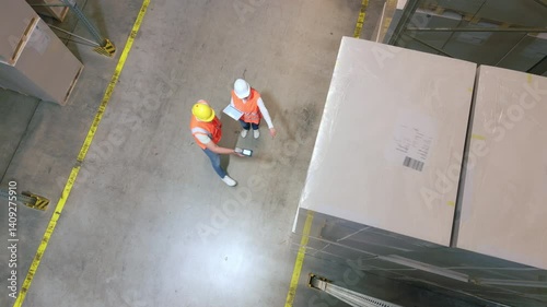 A bird's-eye view of two workers in a warehouse, inspecting packages near a yellow line, emphasizes logistics and organization.