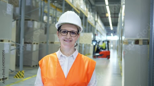 A woman in a hard hat and orange vest stands confidently in a warehouse, overseeing the organized storage of cargo.