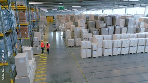 Aerial view of a vast warehouse filled with stacked cargo boxes and two workers in orange vests walking down the aisle.