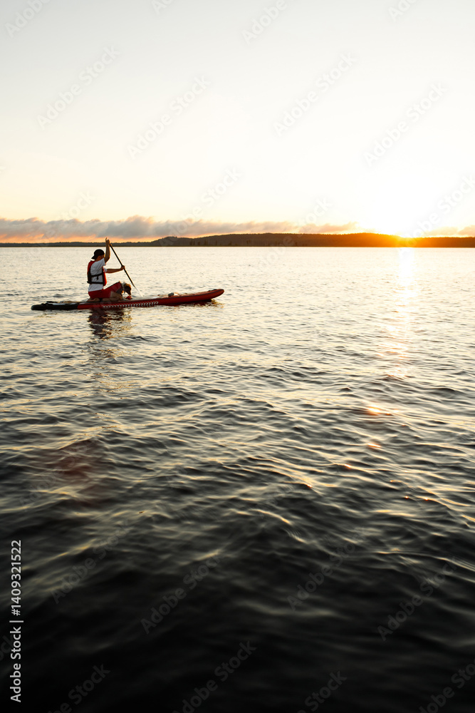 Fototapeta premium Border Collie puppy in red life jacket for animals swimming on a river on a red SUP board along with his owner in a white t-shirt, red shorts and life jacket who holds a paddle on a morning dawn