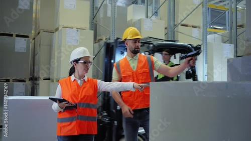 In a warehouse, two workers in orange vests and hard hats collaborate, with one using a tablet and the other holding a tool. The scene highlights teamwork and logistics management.
