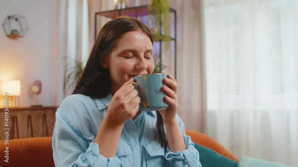 Beautiful smiling happy Caucasian young woman drinking a cup of warm coffee or herbal tea sitting on home orange couch in the morning. Girl enjoying comfortable relaxing alone at living room apartment Beautiful smiling happy Caucasian young woman drinking a cup of warm coffee or herbal tea sitting on home orange couch in the morning. Girl enjoying comfortable relaxing alone at living room apartment
