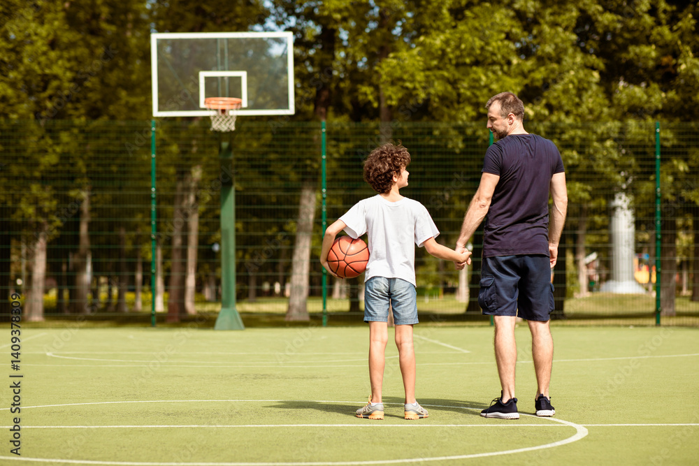 Obraz premium Back view full length portrait of mature man and boy standing on sports field with basket ball, holding hands
