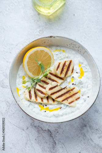 Bowl of greek tzatziki or cucumber yogurt sauce with grilled halloumi cheese, vertical shot on a white stone background, elevated view