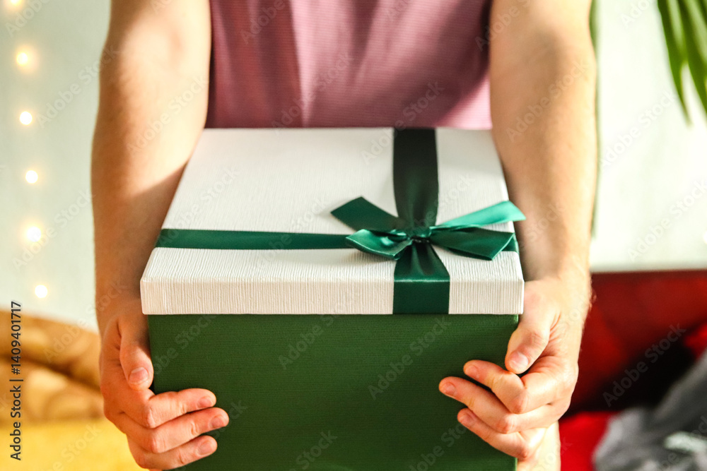 A man hand holding a green gift box with a ribbon, isolated on a neutral background. The composition is clean and minimal, emphasizing the concept of giving, celebration, or a surprise