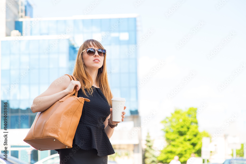 Fototapeta premium Urban woman commuter walking drinking coffee. Young adult woman takeaway coffee in hands. Businesswoman in the city street drinking morning coffee in sunglasses.