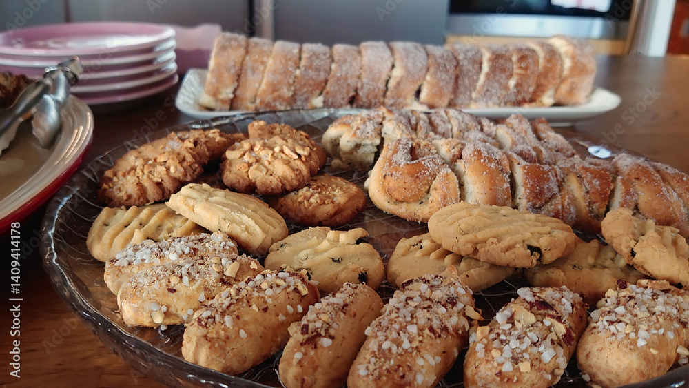 Close-up of a dessert table featuring assorted cookies and pastries, including sugar-dusted braided loaves and nut-topped cookies, arranged on glass platters beside stacked pink plates. Close-up of a dessert table featuring assorted cookies and pastries, including sugar-dusted braided loaves and nut-topped cookies, arranged on glass platters beside stacked pink plates.