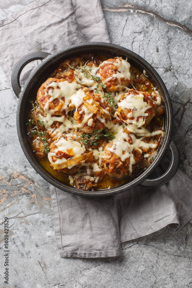 French onion meatballs are nestled into a rich, savory onion sauce and topped with Gruyere and mozzarella cheese closeup on the pan on the table. Vertical top view from above