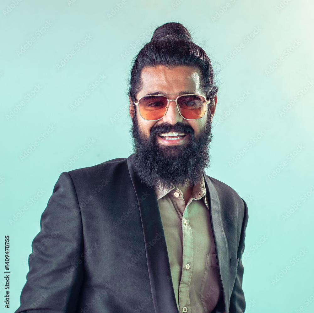Fototapeta premium smiling man with long dark hair tied in a topknot, a full beard, and sunglasses. He is wearing a dark suit jacket over a light-colored collared shirt, standing against a pale teal background.