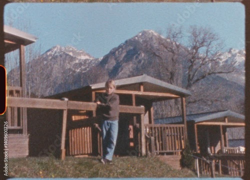Vintage, slightly shaky footage shows a blond girl standing in front of a wooden fence in a mountain campsite, with snowy peaks in the background