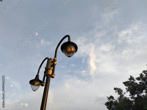 Two vintage-style streetlights against a bright, partly cloudy sky. Urban or park lighting fixture