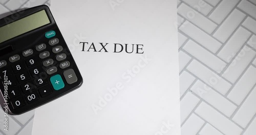 A black calculator rests on a white sheet of paper boldly labeled “TAX DUE,” set against a herringbone-patterned surface.