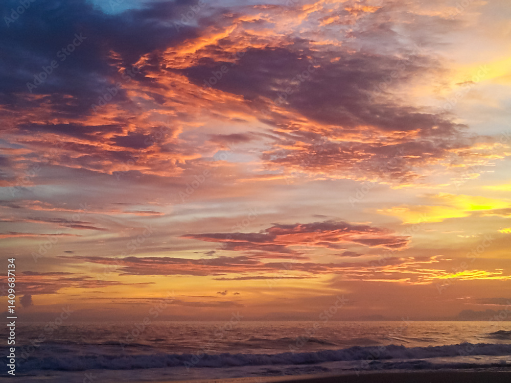 Brilliant sun dips below the ocean horizon, painting the sky with vibrant yellow and orange hues. Its reflection shimmers on the gentle waves washing onto dark sand beach along the Costa Rican coast