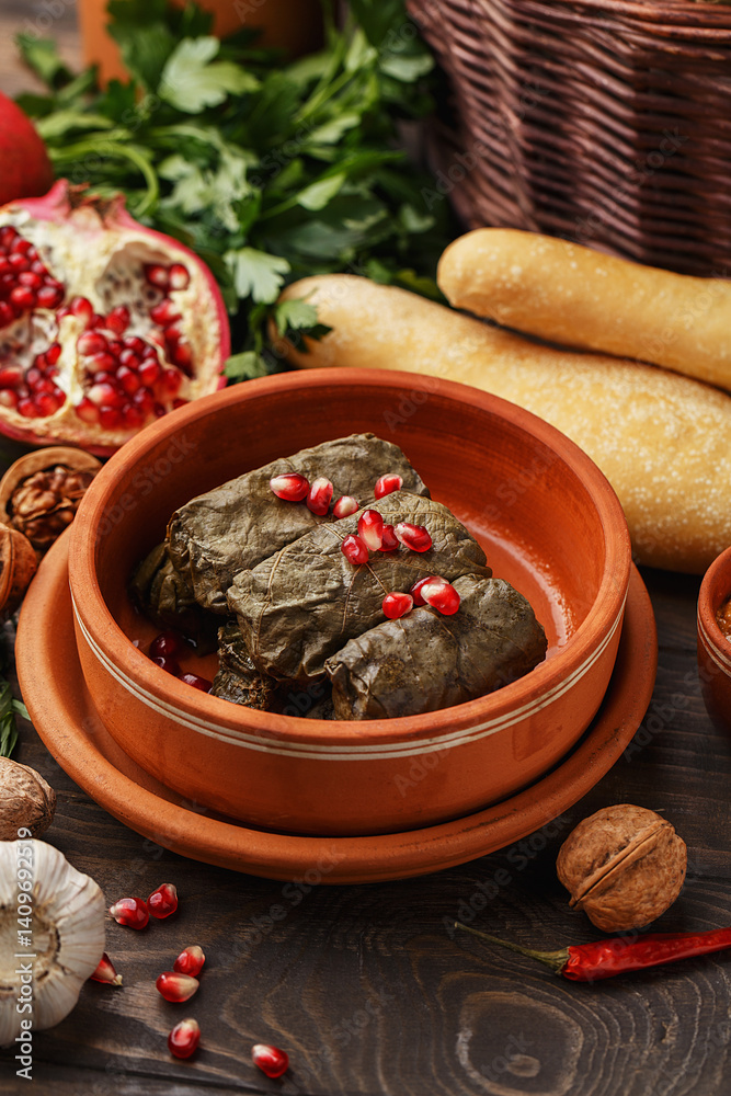 Fototapeta premium Traditional middle eastern dish dolma. Grape leaves stuffed with rice and meat. Tolma or sarma served in clay bowl decorated with pomegranate seeds over wooden table. Close up view