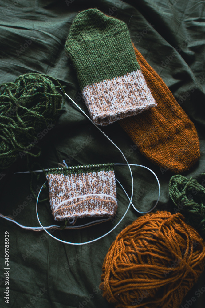 Dark green and brown yarn ball with knitted socks and knitting needles. Wool threads with deep green background. Woven crochet close up. Knitting hobby. Knitwear concept. Needlework background.