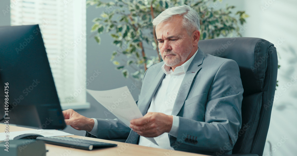An experienced businessman, grey-haired man head of company is sitting at desk in a comfortable chair of the president, looking at documents brought to him, reading them carefully, asking a colleague An experienced businessman, grey-haired man head of company is sitting at desk in a comfortable chair of the president, looking at documents brought to him, reading them carefully, asking a colleague
