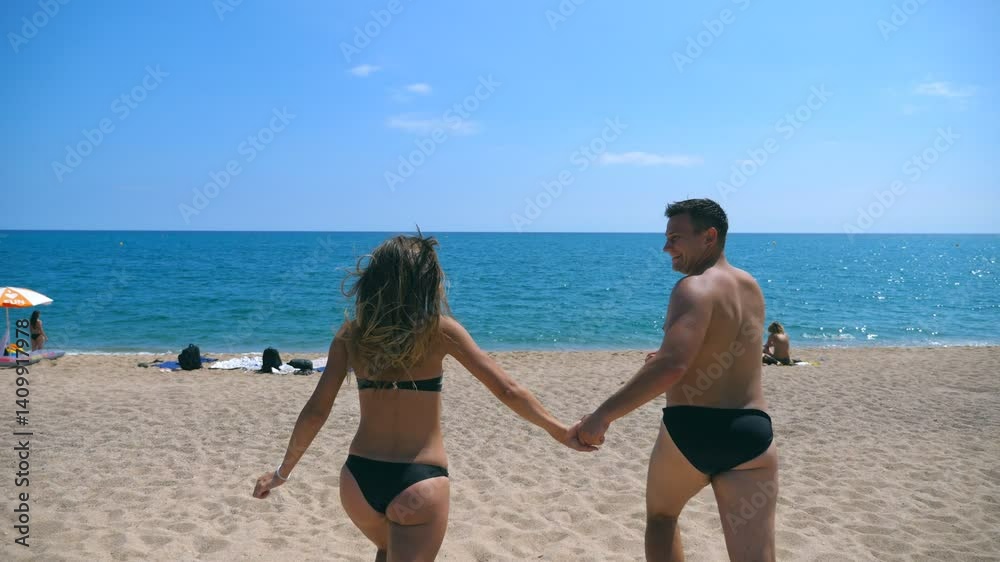 Happy couple holds hands of each other running on sandy beach to the sea. Young smiling lovers jogging to seaside at sunny day. Cheerful man and woman having fun together at seashore. Vacation concept Happy couple holds hands of each other running on sandy beach to the sea. Young smiling lovers jogging to seaside at sunny day. Cheerful man and woman having fun together at seashore. Vacation concept
