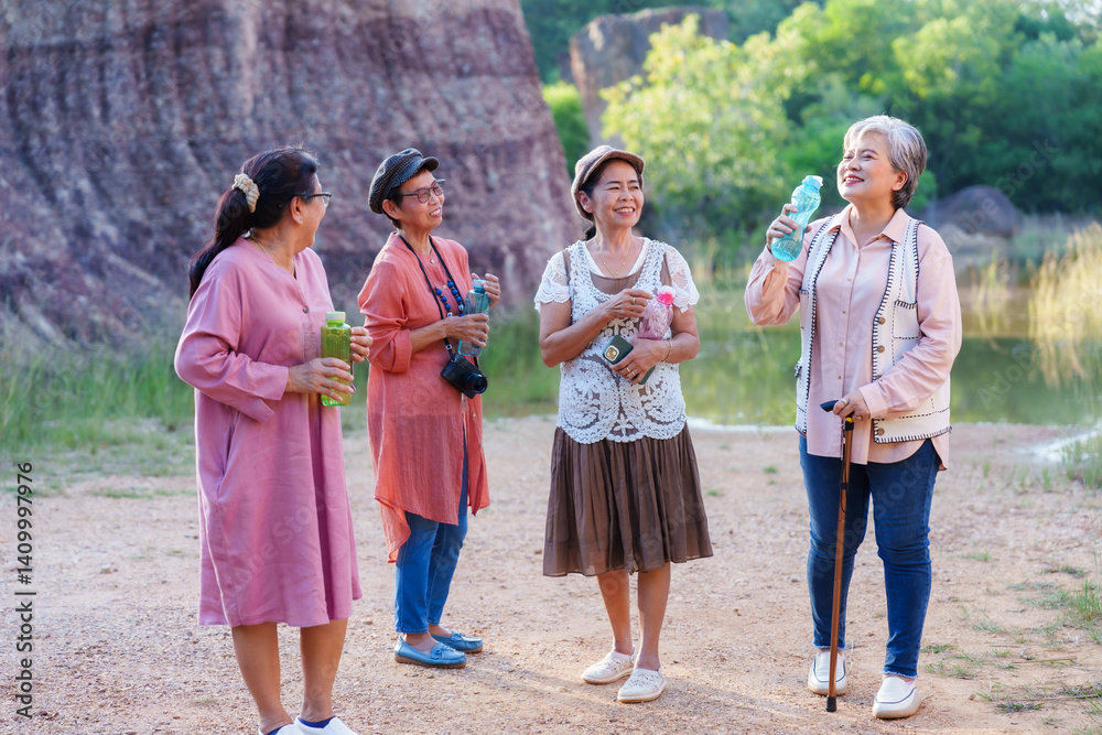 Four senior Asian women enjoying nature trip while standing and drinking water in group, dressed in casual clothing with hats and cane, smiling and relaxing together during peaceful outdoor moment Four senior Asian women enjoying nature trip while standing and drinking water in group, dressed in casual clothing with hats and cane, smiling and relaxing together during peaceful outdoor moment
