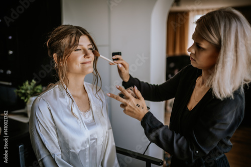 Fényképezés Makeup artist applying blush to smiling bride wearing a silk robe during wedding preparations in a cozy, bright room