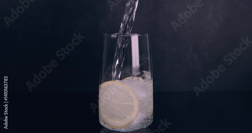 Carbonated mineral water flowing into a glass with lemon slices on black background. Healthy and fresh water. Slow motion