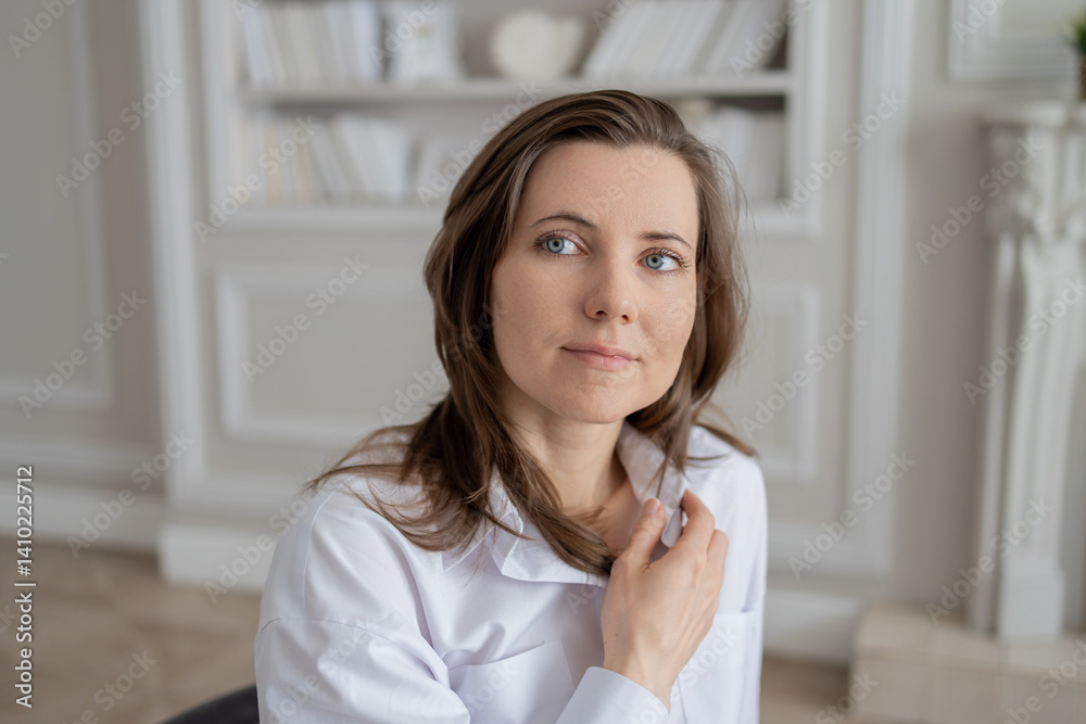 a young woman with brown hair and blue eyes, dressed in a white shirt. She is sitting on a chair in a bright room with a bookcase in the background. He looks directly at the camera, lightly touching h a young woman with brown hair and blue eyes, dressed in a white shirt. She is sitting on a chair in a bright room with a bookcase in the background. He looks directly at the camera, lightly touching h