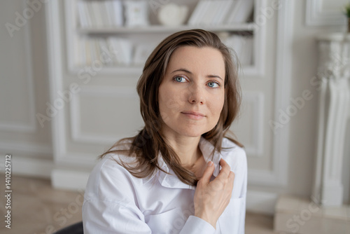 a young woman with brown hair and blue eyes, dressed in a white shirt. She is sitting on a chair in a bright room with a bookcase in the background. He looks directly at the camera, lightly touching h Free Download a young woman with brown hair and blue eyes, dressed in a white shirt. She is sitting on a chair in a bright room with a bookcase in the background. He looks directly at the camera, lightly touching h - GFXLoad